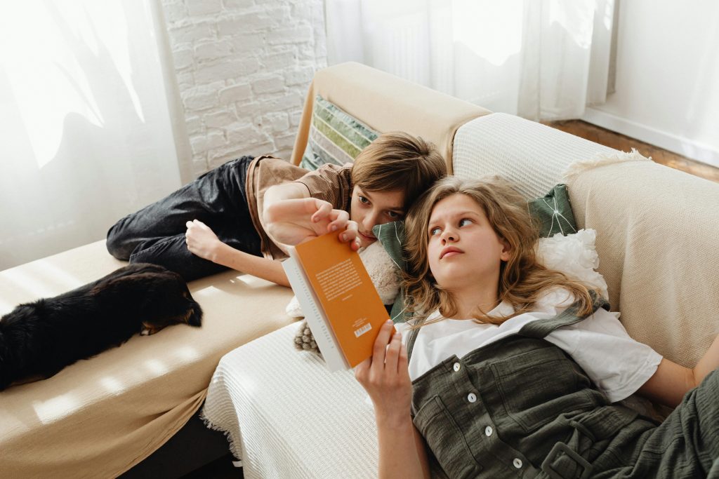 Two teenage siblings relax on a couch reading a book together in a cozy indoor setting.
